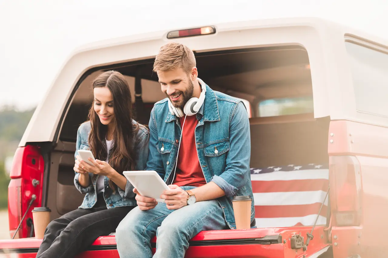 Young Couple Using Gadgets While Sitting in Car Trunk During Trip